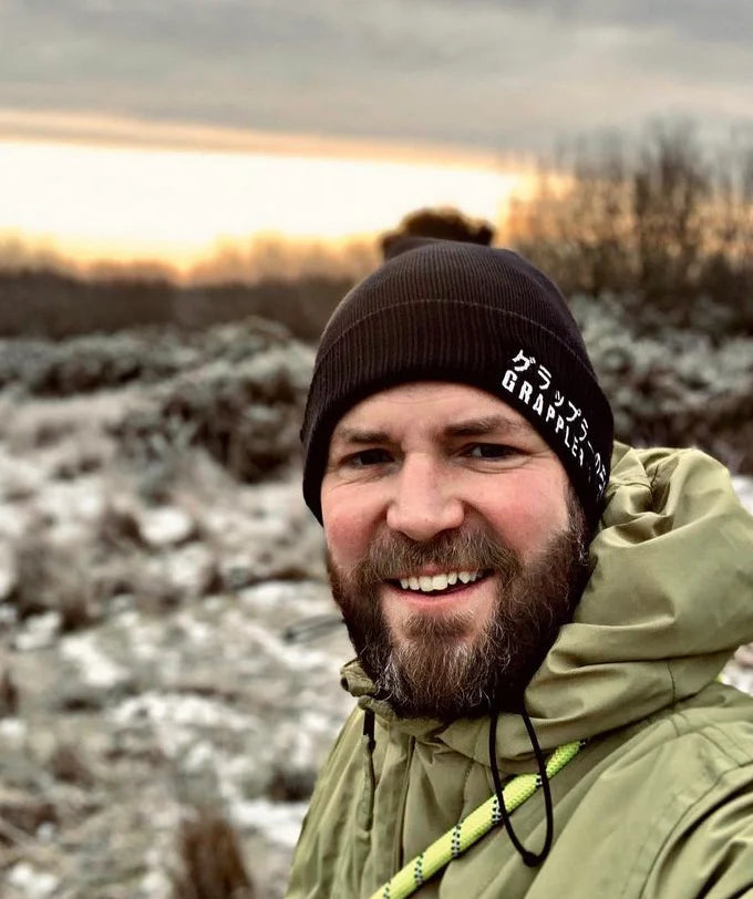 A man wearing sunglasses with a light beard smiles outdoors.